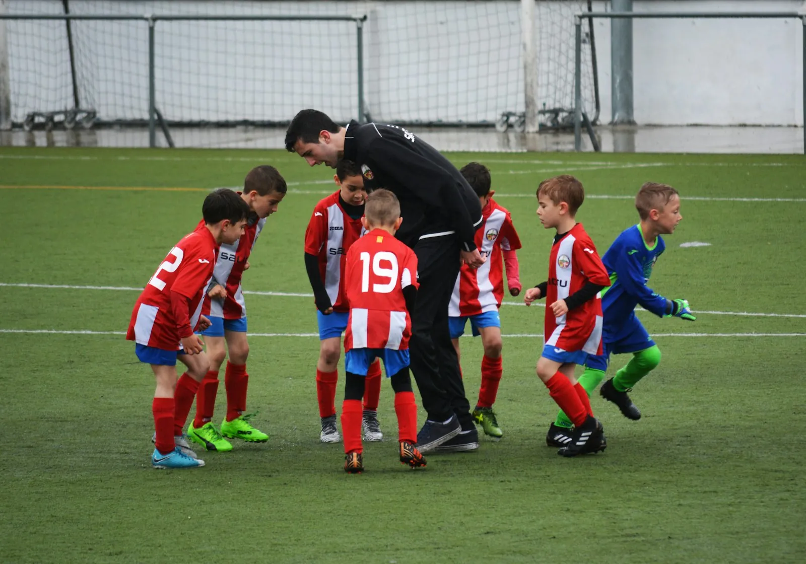 A football coach talking to a group of young players in red kits on a training pitch