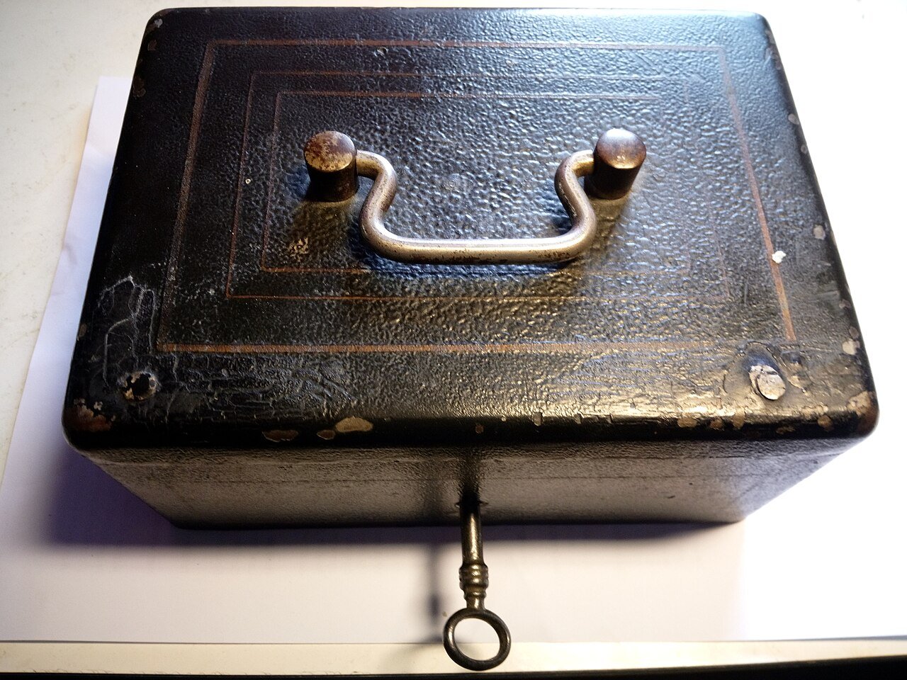 An old black metal money box with a carry handle on the lid and a key in the lock