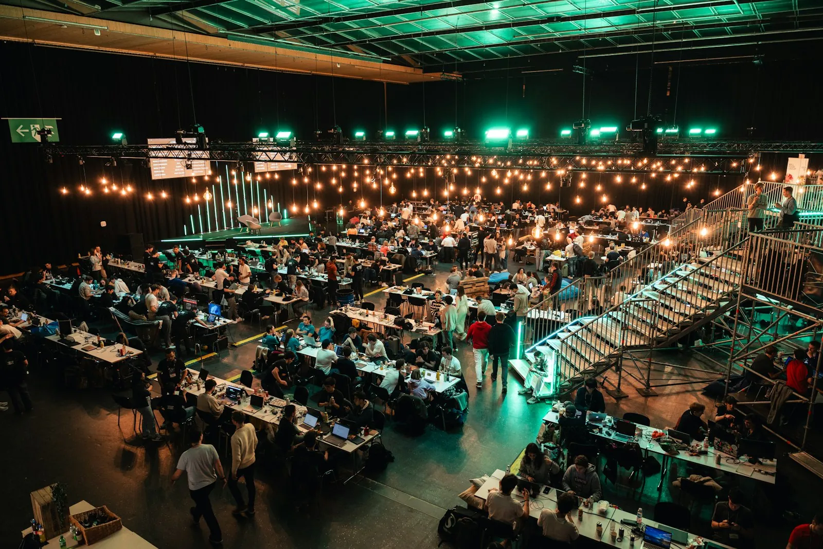 An overhead view of a huge hackathon hall packed with teams coding at long tables under strings of lights
