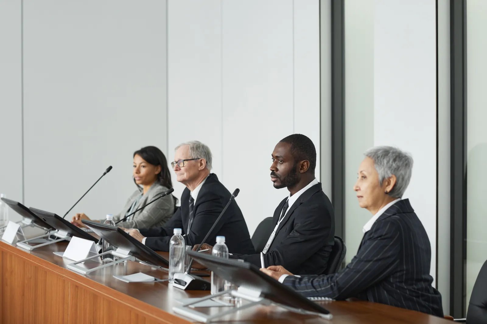 A four-member panel sits behind a long table with microphones and screens, listening closely