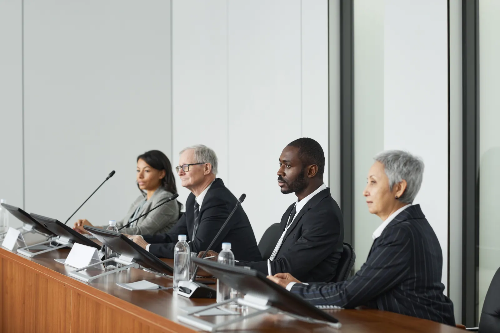 A four-member panel sits behind a long table with microphones and screens, listening closely