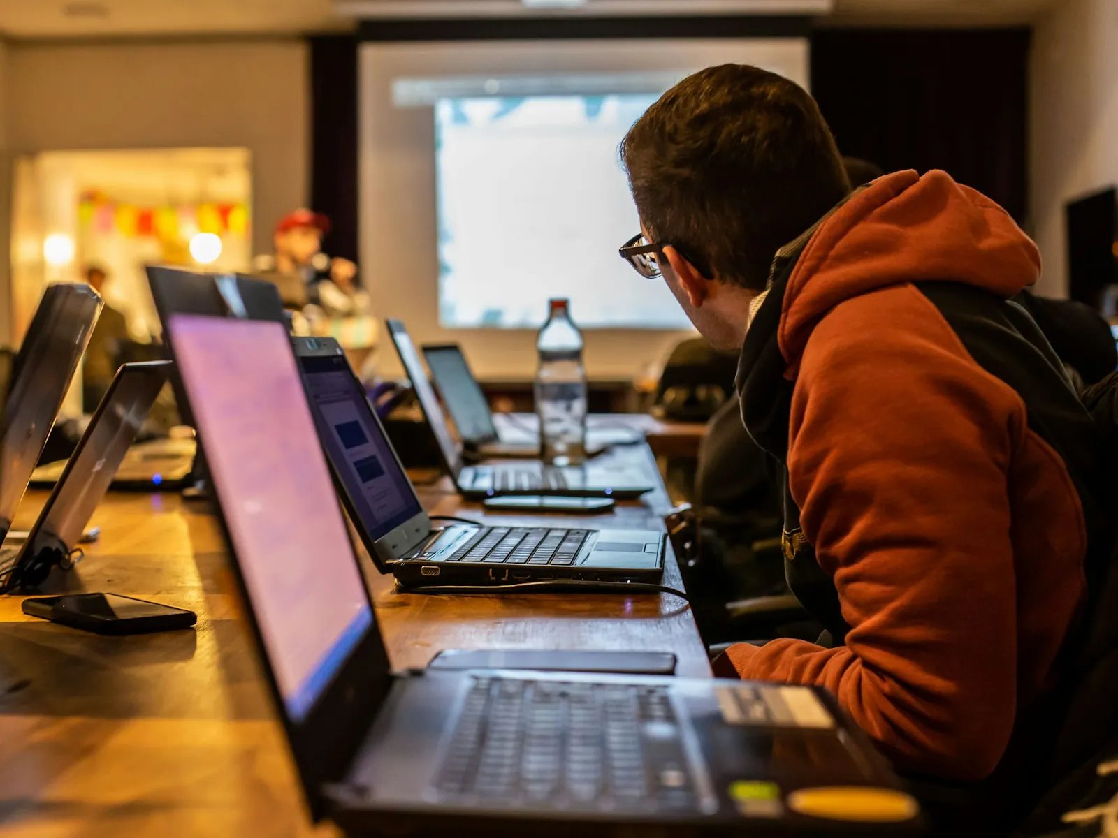 A participant in an orange hoodie leans over a row of glowing laptops in a dim room during a long coding session