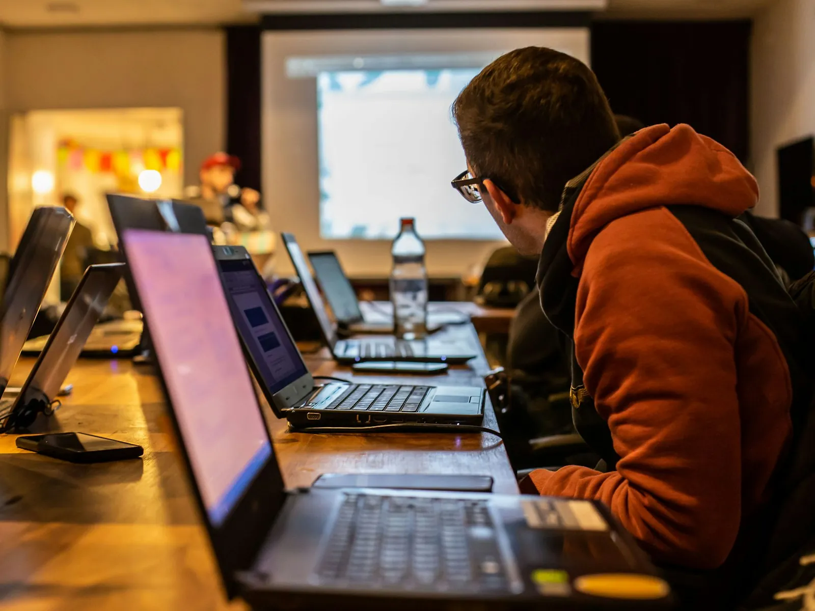 A participant in an orange hoodie leans over a row of glowing laptops in a dim room during a long coding session