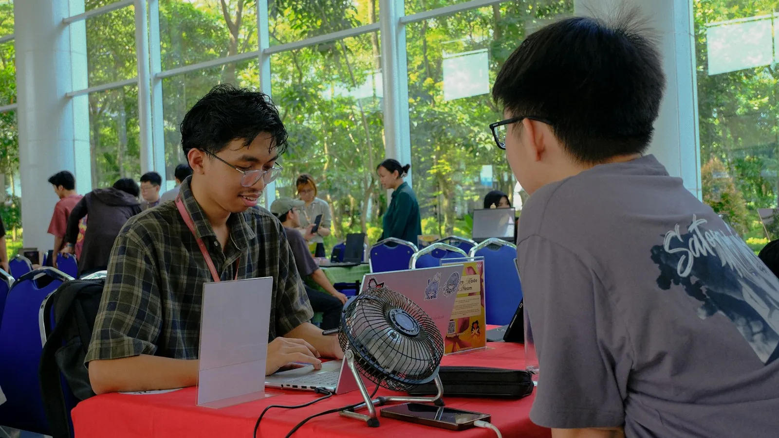 A student demos a project on a laptop to a visitor across an exhibition table at a campus tech event
