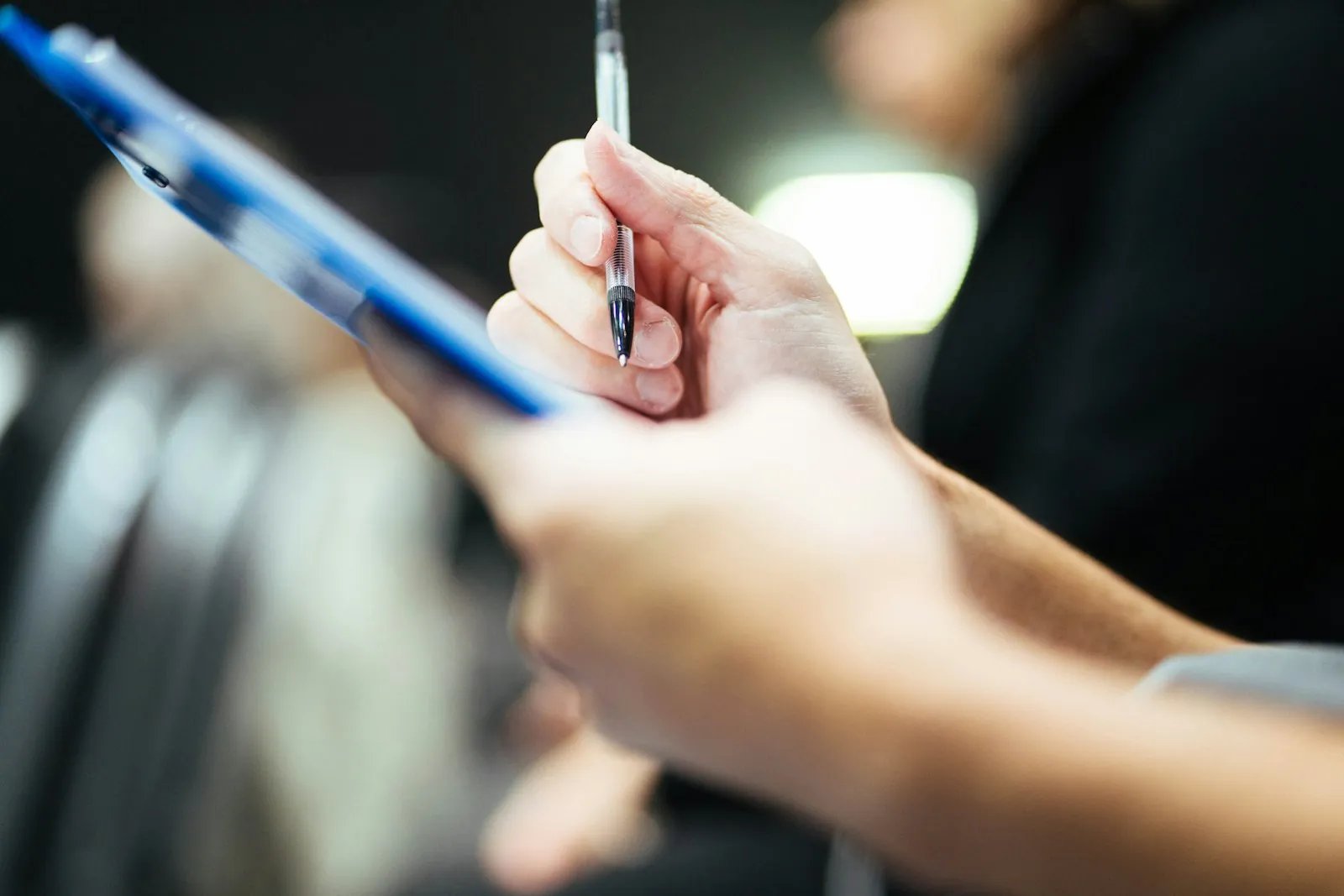 An evaluator holds a pen over a blue clipboard, ready to write down scores