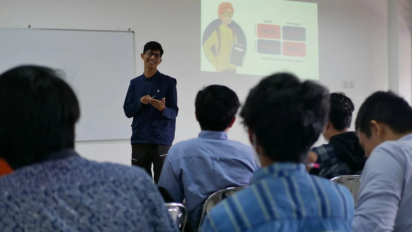 A smiling student presents in front of a projected slide while a room of fellow students listens