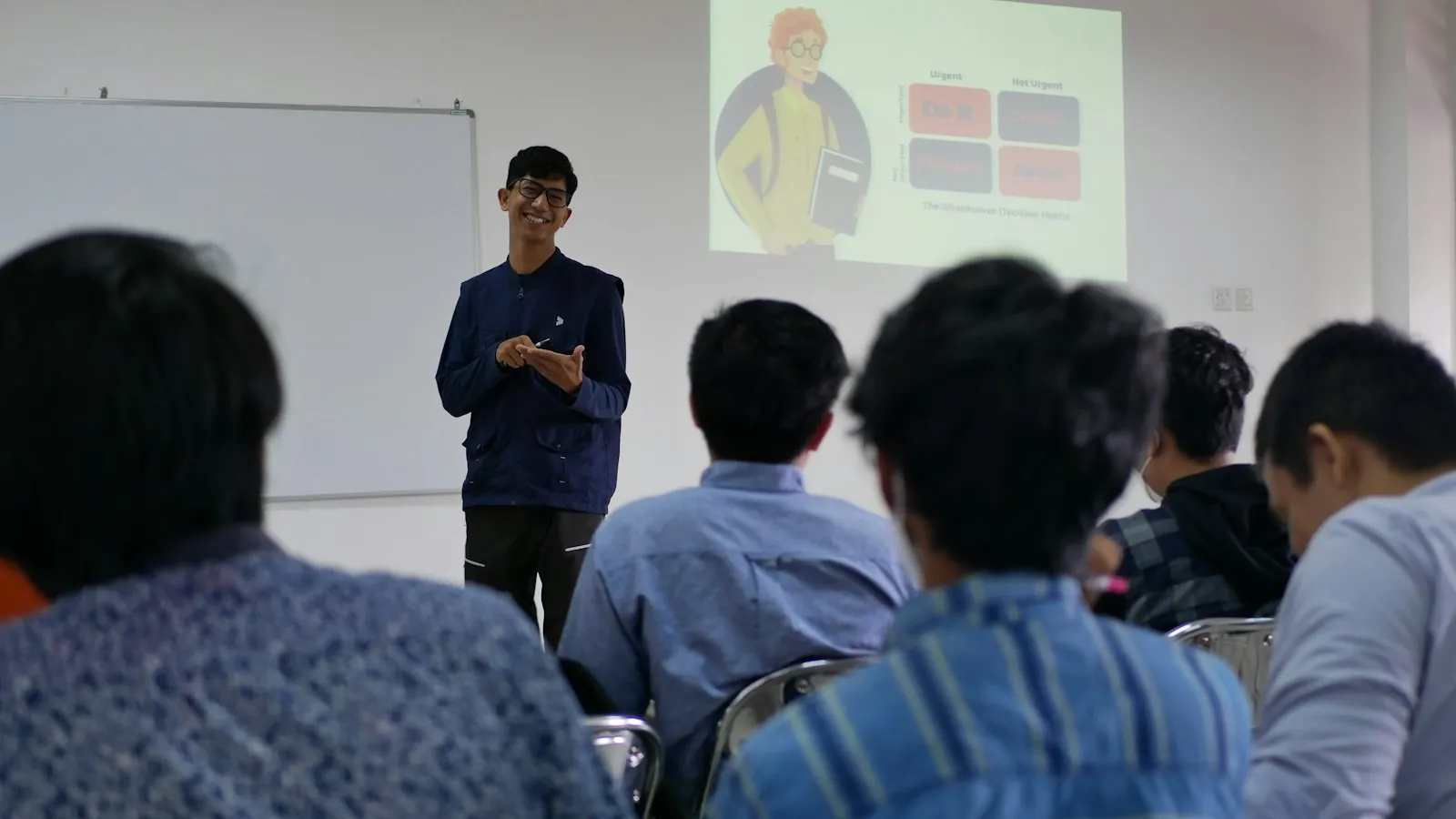 A smiling student presents in front of a projected slide while a room of fellow students listens