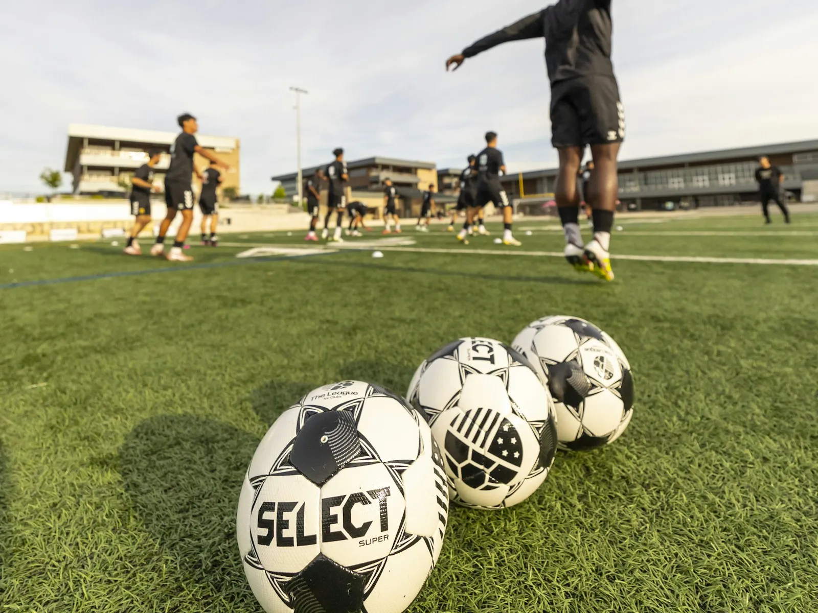 Three footballs lined up on artificial grass while a squad in black kit warms up on the pitch behind them