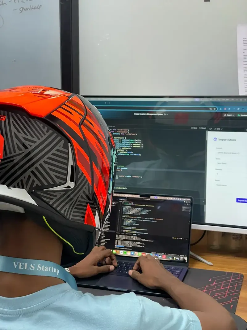 Rahul in a motorbike helmet, coding on a MacBook in front of a large monitor