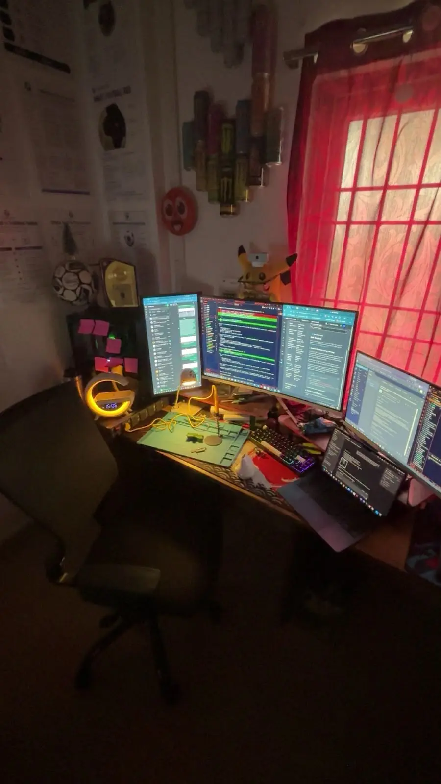 Rahul's desk at night: three monitors, a laptop, sticky notes and a wall of Monster cans