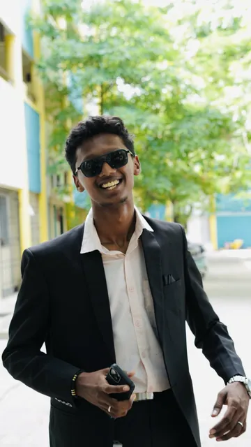 Rahul Dhileep Kumar in a black suit and sunglasses, laughing on a tree-lined street